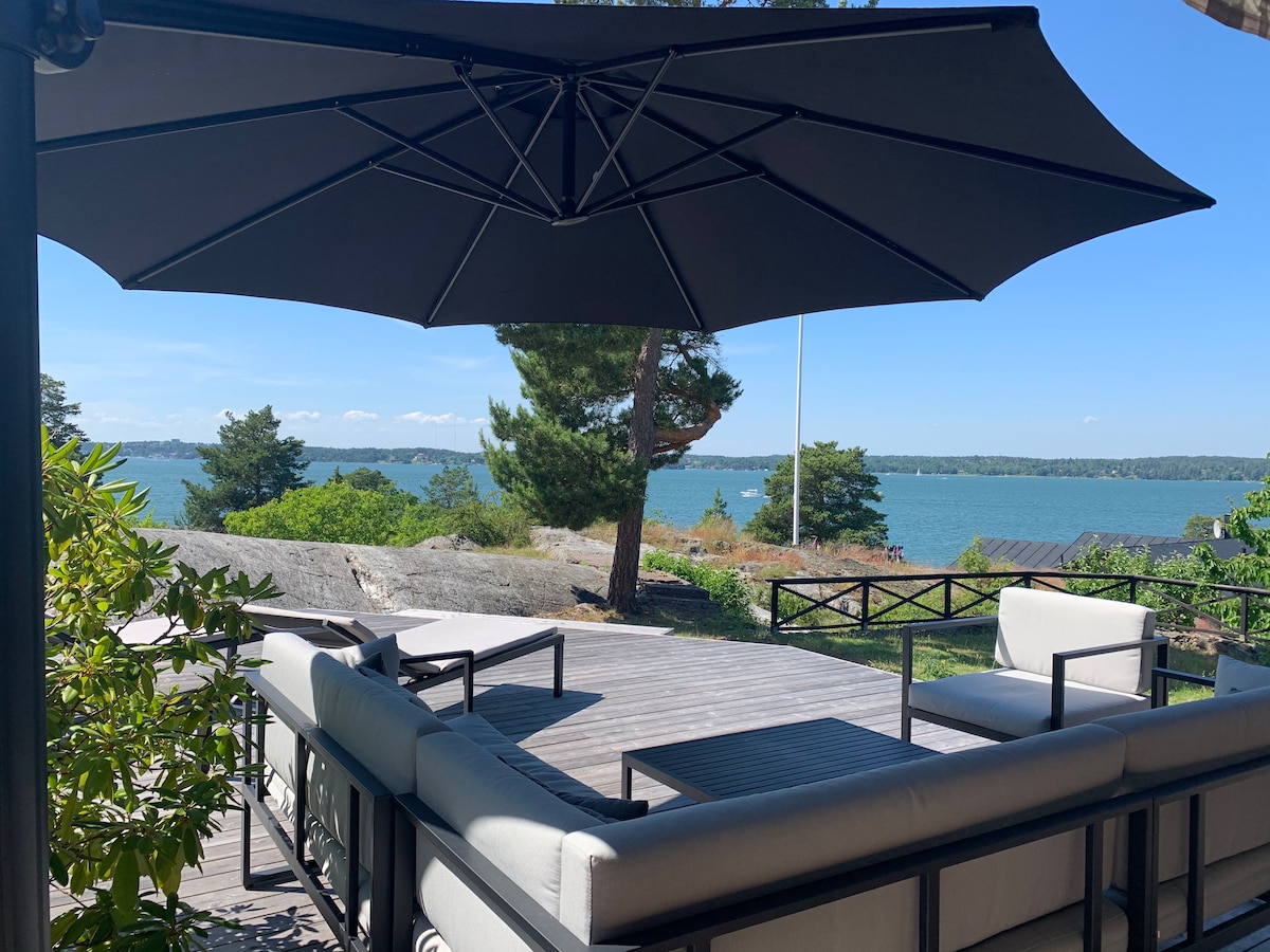 An outdoor lounge area is situated under a large umbrella, featuring comfortable seating and a coffee table. A view of the serene waterfront and surrounding greenery is visible in the background, creating a relaxing atmosphere for outdoor gatherings.