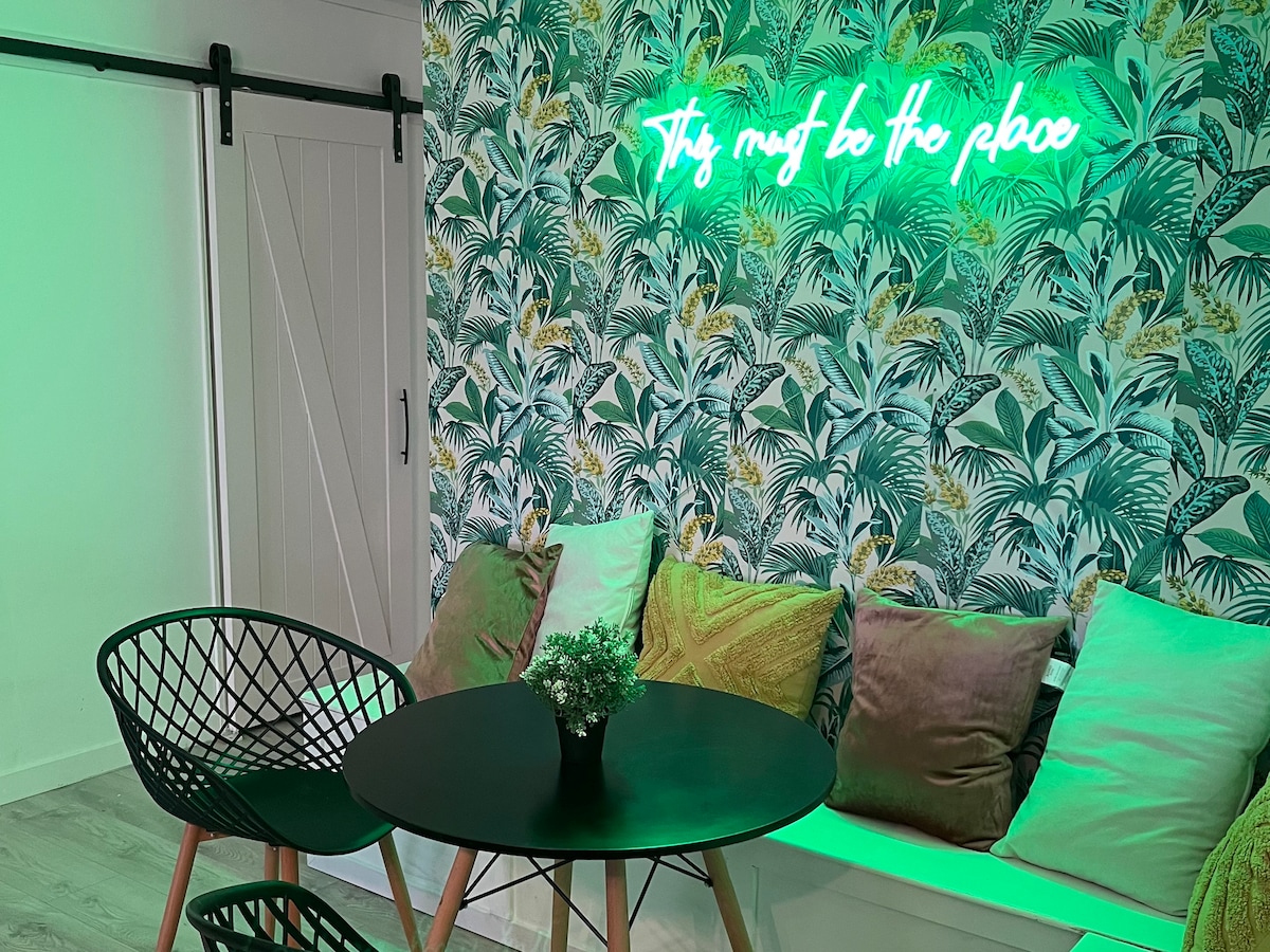 A cozy sitting area features a round black table surrounded by two stylish chairs. A mural with lush green foliage adds color to the wall. Soft cushions in various textures are placed on a built-in bench, illuminated by a glowing neon sign above.
