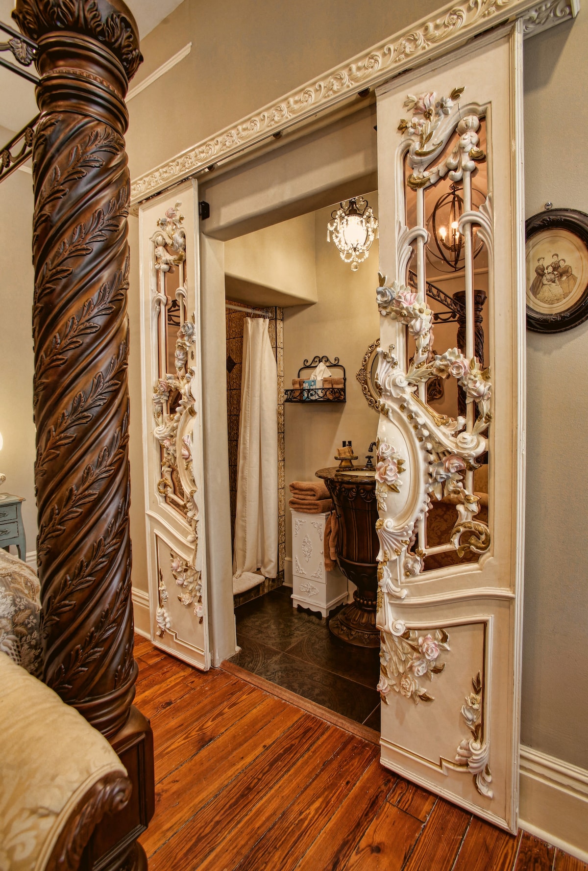 An ornately designed doorway with vintage floral carvings leads to a private bathroom. The warm wood floor contrasts with the intricately detailed door, enhancing the space's character. Soft lighting from a nearby fixture illuminates the entrance, creating a welcoming transition from the bedroom.