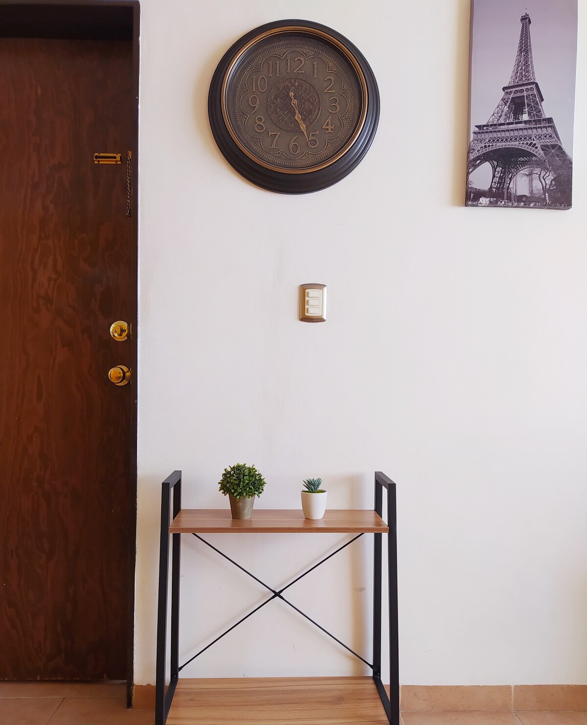 An entrance area is featured with a wooden door, adorned with a round wall clock and black-and-white artwork depicting the Eiffel Tower. A small, minimalist table holds two potted plants, enhancing the space's inviting appearance.