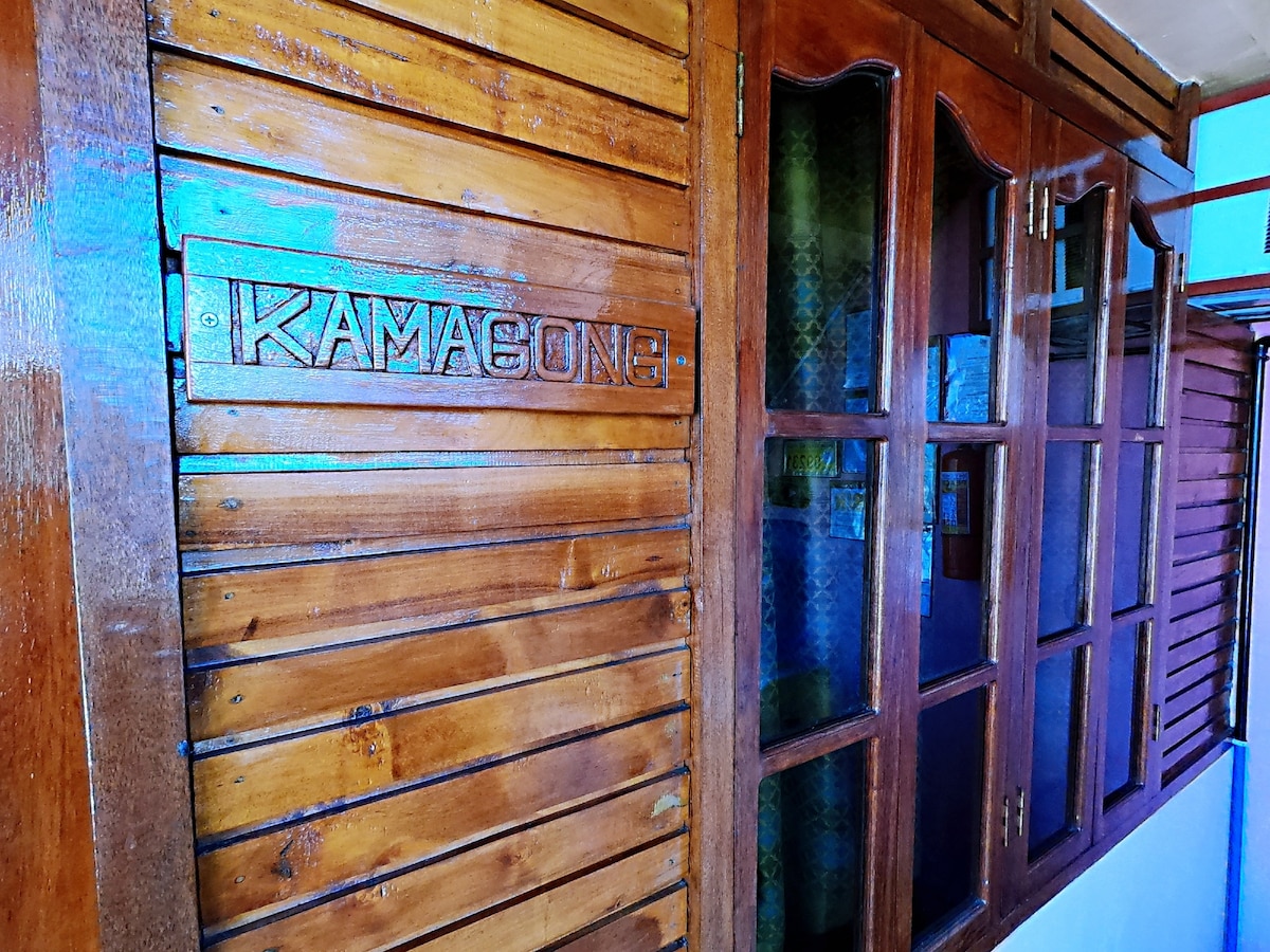 A wooden wall with the name "KAMASONG" carved prominently at the top is featured. The wall includes multiple window frames with transparent glass panes, allowing glimpses of the interior decor. The warm tones of the wood enhance the natural aesthetic.