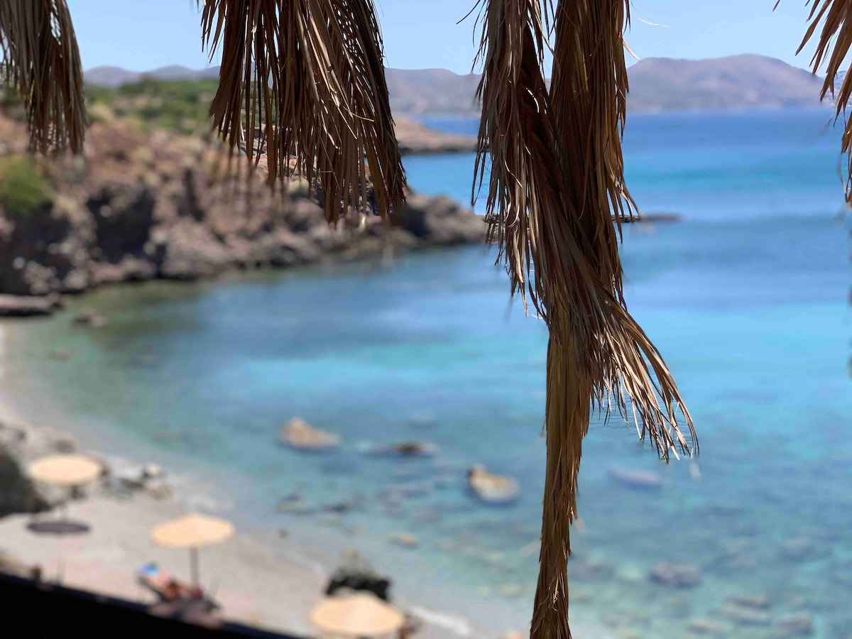 A serene beach scene is captured, featuring clear turquoise waters lapping against a sandy shore. Sun loungers with umbrellas are positioned on the beach, while rocky formations are visible in the background. The image is framed by palm fronds, adding a natural touch.