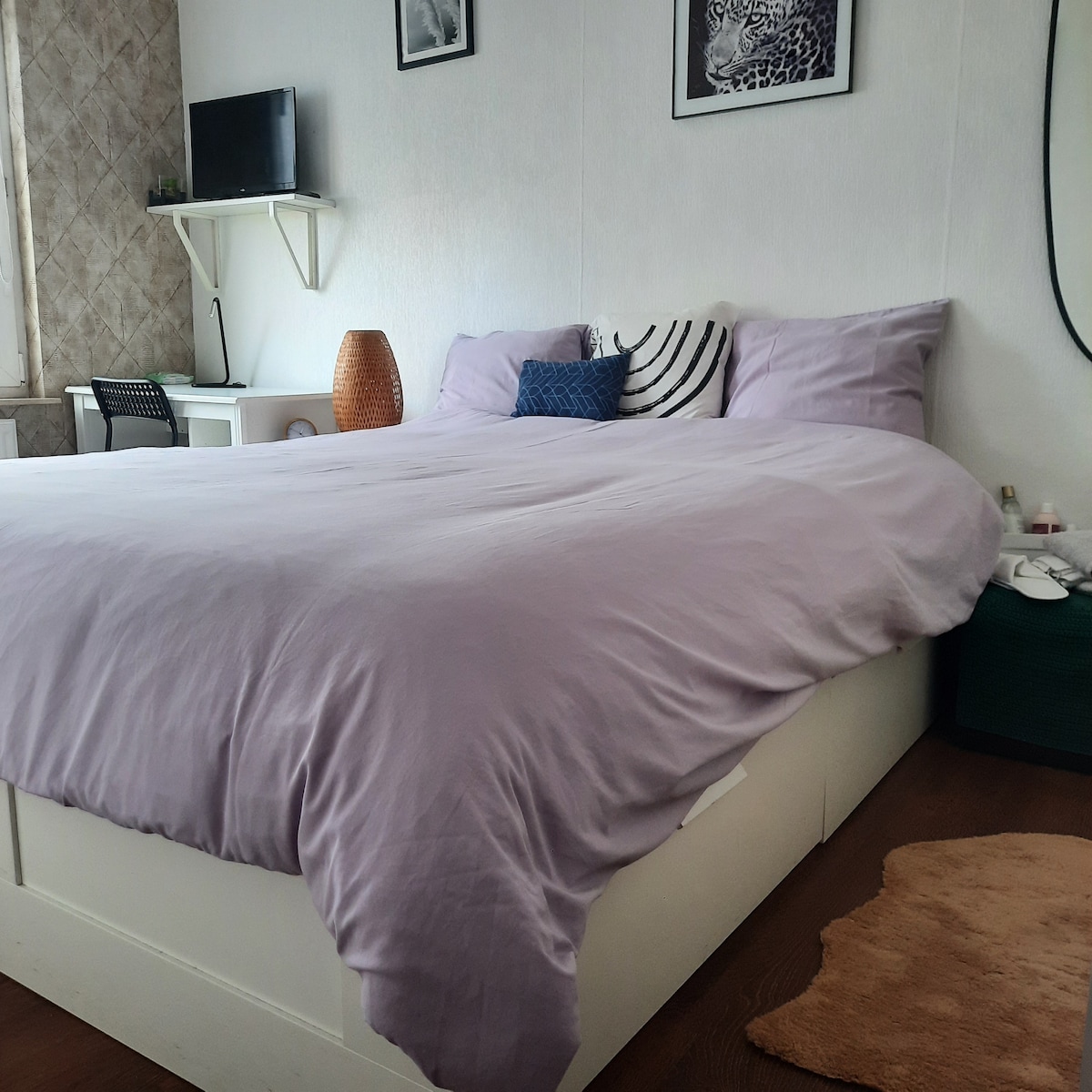 A spacious bed is covered with a light purple comforter, complemented by decorative pillows. A small desk and a chair are visible in the background, with a television mounted on the wall. Natural light illuminates the room, enhancing the calming atmosphere.