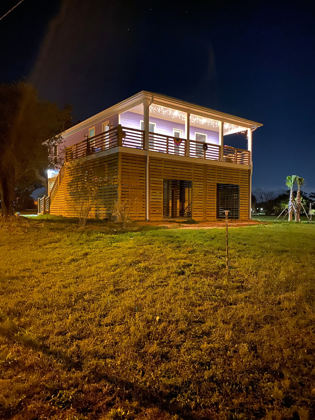 A modern two-story house is illuminated at night, showcasing a wooden exterior with slatted design. The balcony is adorned with string lights, creating a welcoming ambiance. The surrounding yard features subtle landscaping, with grass extending toward the distant tree line.