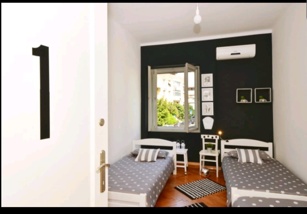 A room with two twin beds is featured, each adorned with simple bedding. A window with a decorative black frame allows natural light to enter. An air conditioning unit is mounted on the wall, and minimalistic artwork is arranged above a small chair and table.