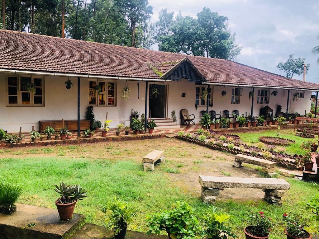 A single-story bungalow is surrounded by well-maintained gardens, featuring potted plants and neatly arranged flower beds. The exterior showcases a tiled roof, and seating areas with wooden chairs can be seen along the pathway. Lush trees rise in the background, enhancing the natural setting.