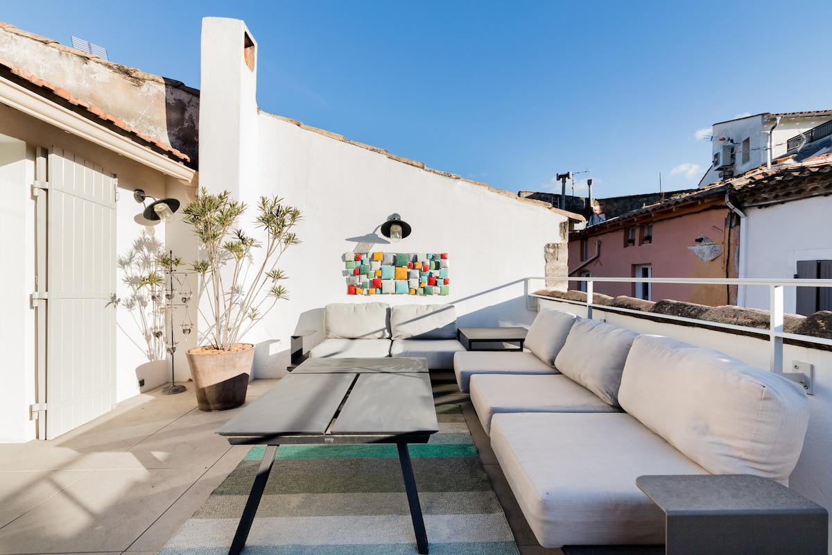 A spacious south-facing terrace features modern seating arrangements, complemented by a decorative wall piece. A potted plant adds a touch of greenery, while the surrounding rooftops provide a glimpse of the village. Natural light brightens the area, enhancing the outdoor experience.