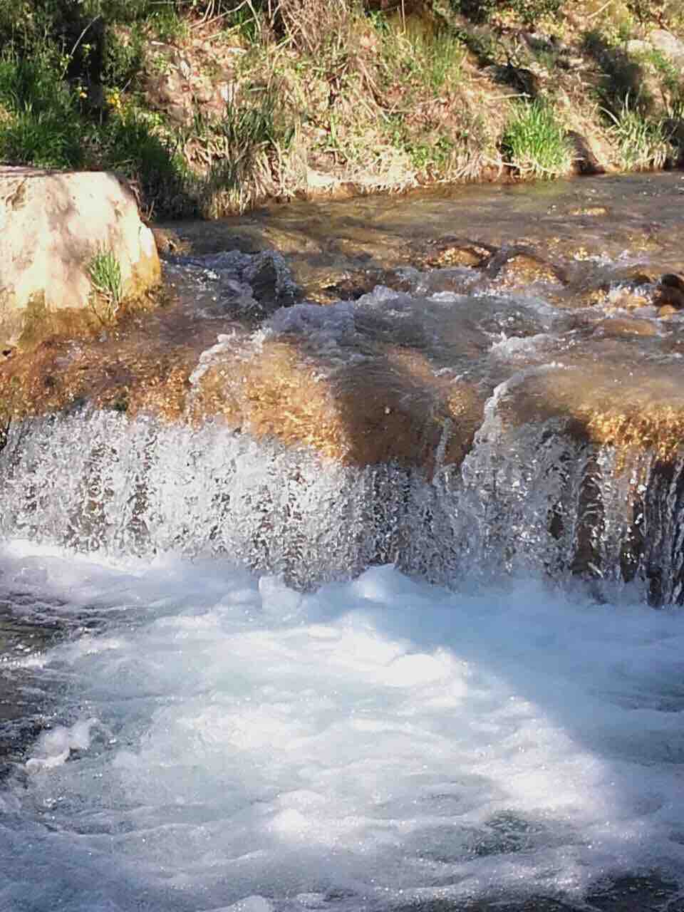 A natural water flow is captured, with clear blue water cascading over smooth stones. The surrounding greenery is visible, enhancing the serene environment. Sunlight creates shimmering reflections on the water's surface, inviting a sense of calmness and connection to nature.