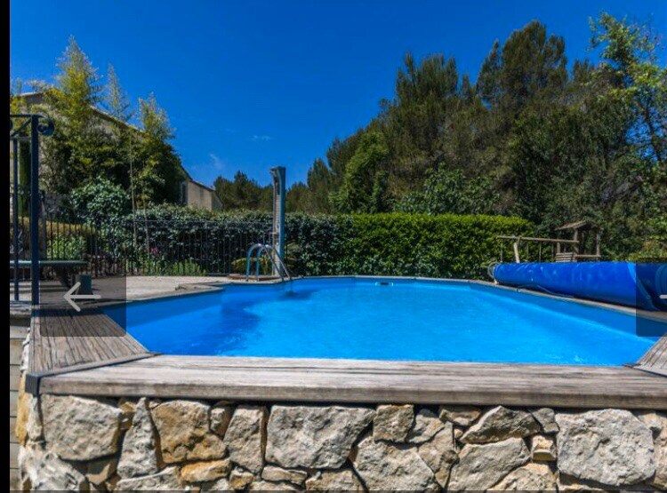 A private swimming pool with clear blue water is surrounded by natural stone walls and wooden decking. Lush greenery and trees provide a serene backdrop, while a slide and steps are visible at one end. The pool area offers a peaceful retreat in a sunlit setting.
