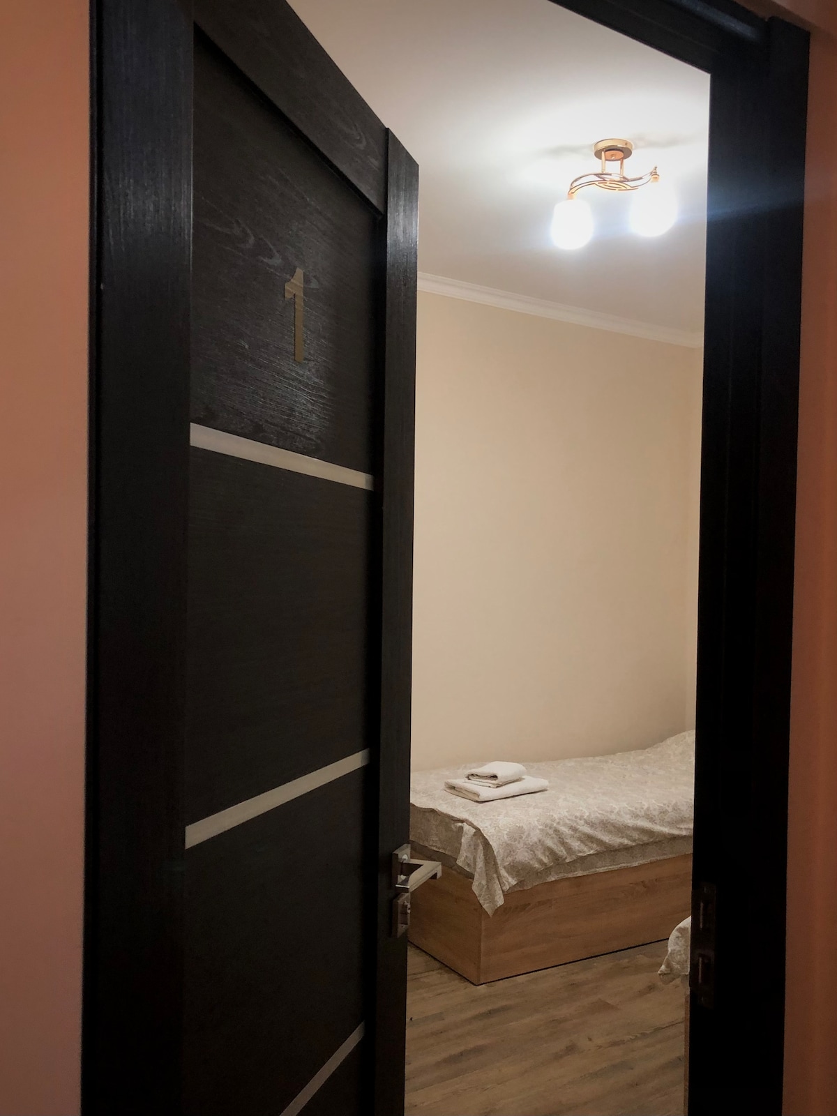 A doorway provides a glimpse into a simple bedroom featuring a spacious bed covered with a light-colored blanket. Soft lighting is provided by a ceiling fixture, and a neat arrangement of towels is placed on the bed.