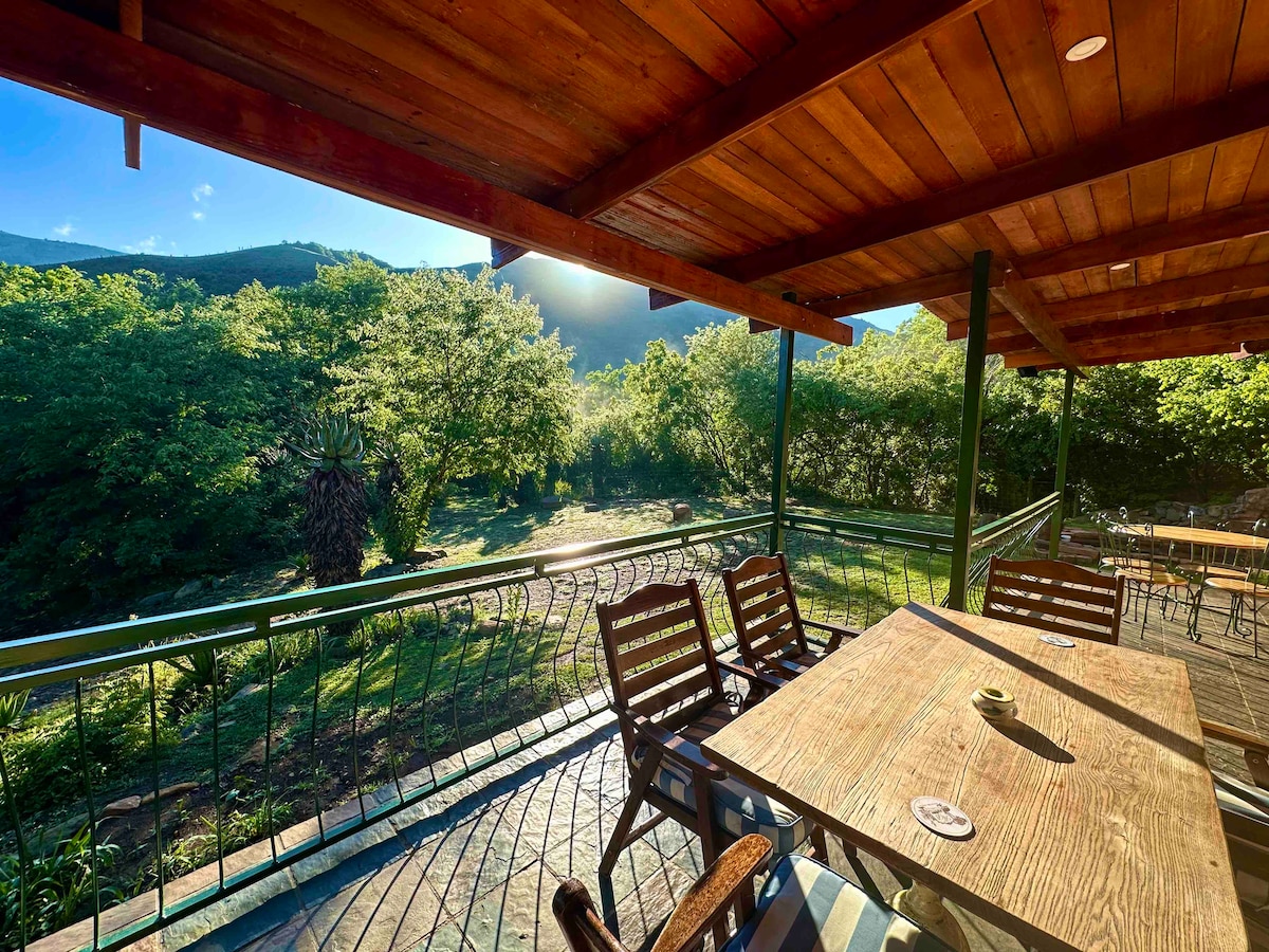A spacious outdoor deck features wooden seating arrangements and a table, overlooking lush greenery and natural landscape. Sunlight gently illuminates the area, creating a serene setting that invites relaxation amid the surrounding nature.