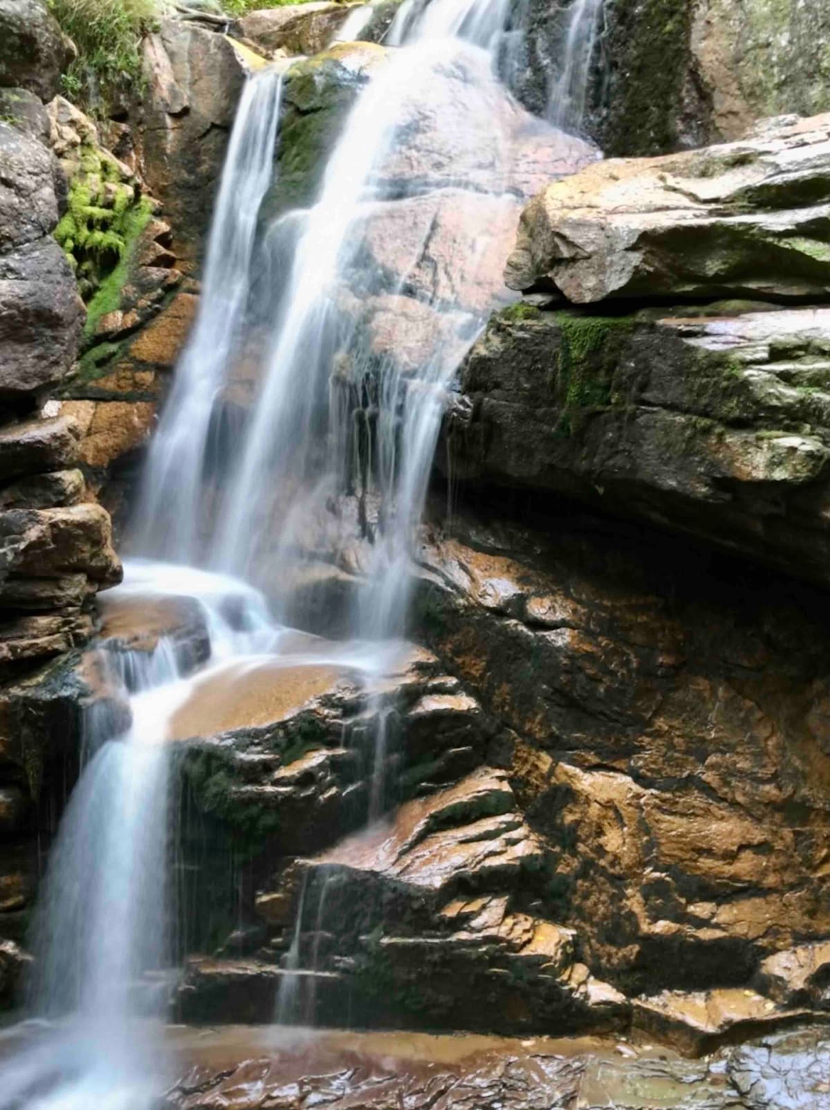 A serene waterfall cascades over rugged stones, surrounded by lush greenery. The crystal-clear water flows down several tiers, creating a tranquil atmosphere. The sunlight reflects off the water, accentuating the natural beauty of the scene.