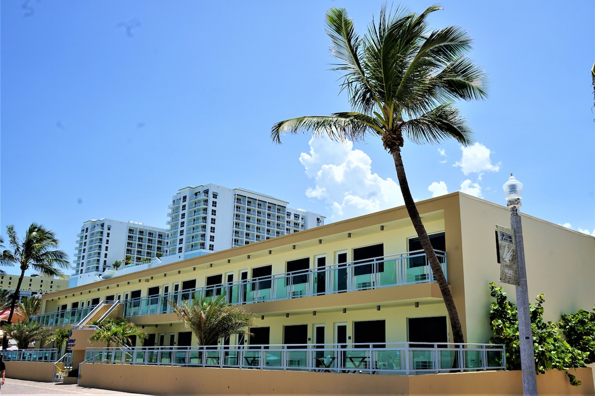 A low-rise building with an inviting yellow exterior features balcony access from each room. Palm trees sway gently in the foreground, complementing the clear blue sky. Nearby, a modern high-rise can be seen, enhancing the location's vibrant atmosphere.