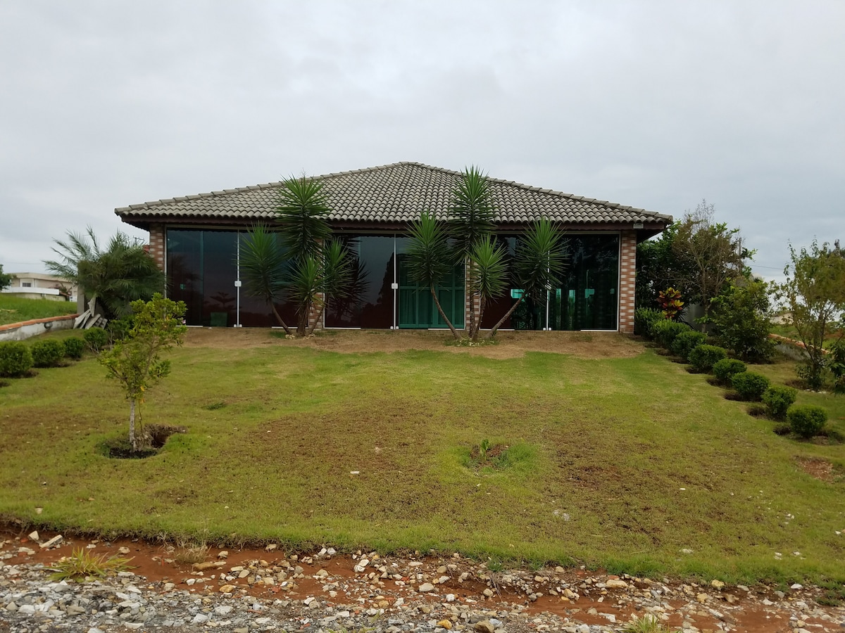 The house features a modern design with a spacious front yard, outlined by well-maintained green shrubs and palm trees. Expansive glass walls invite natural light while providing views of the surrounding area, complemented by a tiled roof.