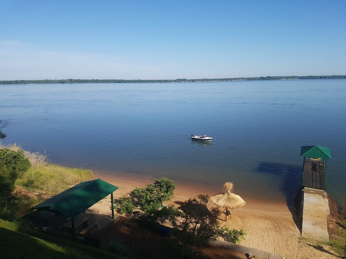 A peaceful riverside scene captures calm waters with a small boat gently drifting. A sandy beach and shaded seating area are visible, framed by lush greenery. The horizon features a clear sky above the tranquil Rio Paraná, enhancing the serene surroundings.