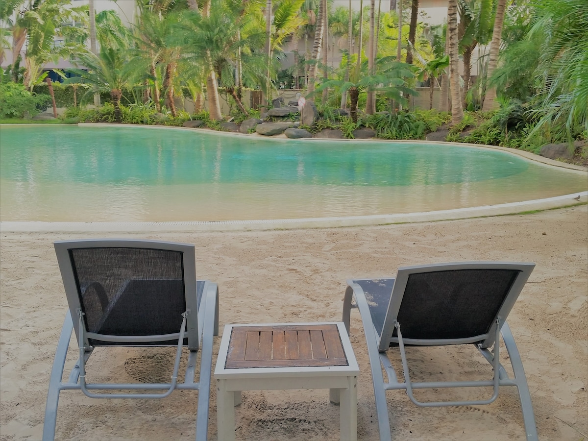 Two lounge chairs are positioned on sandy ground beside a tranquil pool, surrounded by lush tropical greenery. A small wooden table is placed between the chairs, providing a spot for refreshments while enjoying the serene view of the water and the surrounding landscape.