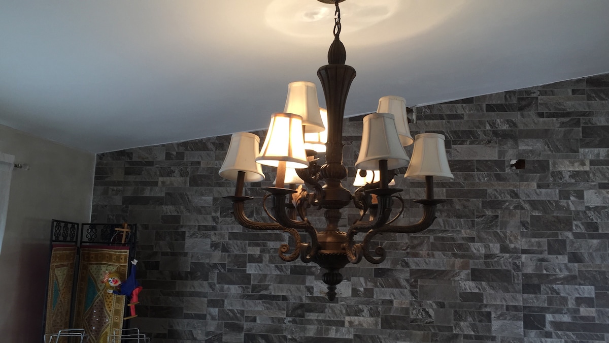 A classic-style chandelier is suspended from the ceiling, featuring multiple arms with cream-colored shades. The wall behind is adorned with textured stone, creating a distinctive backdrop. A small decorative figure can be seen to the left, adding a touch of charm to the space.