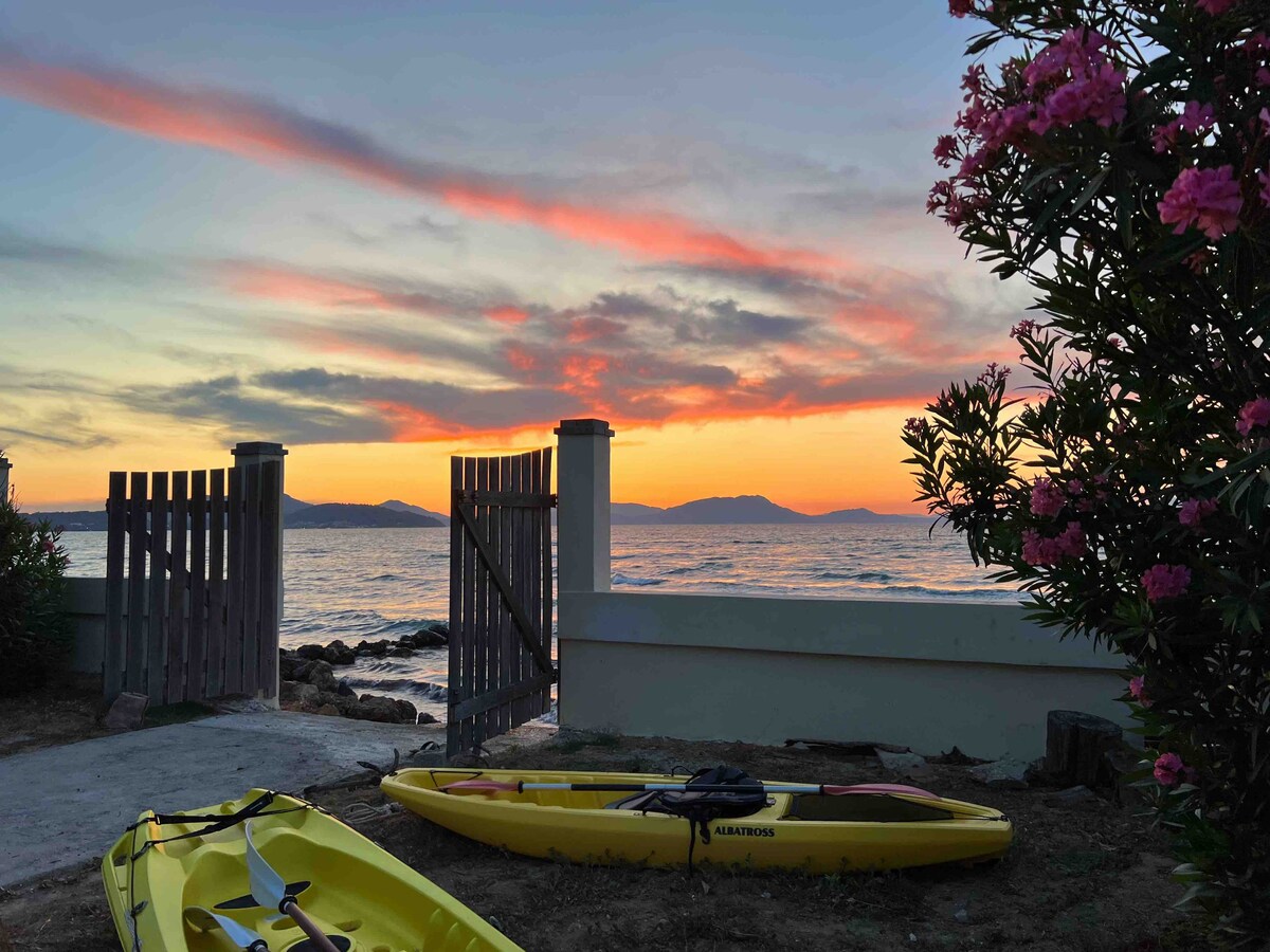 A serene view captures the ocean at sunset, framed by a wooden gate. Kayaks rest on the shore, while vibrant flowers add color to the scene. The sky showcases hues of pink and orange, reflecting a calm atmosphere by the water.