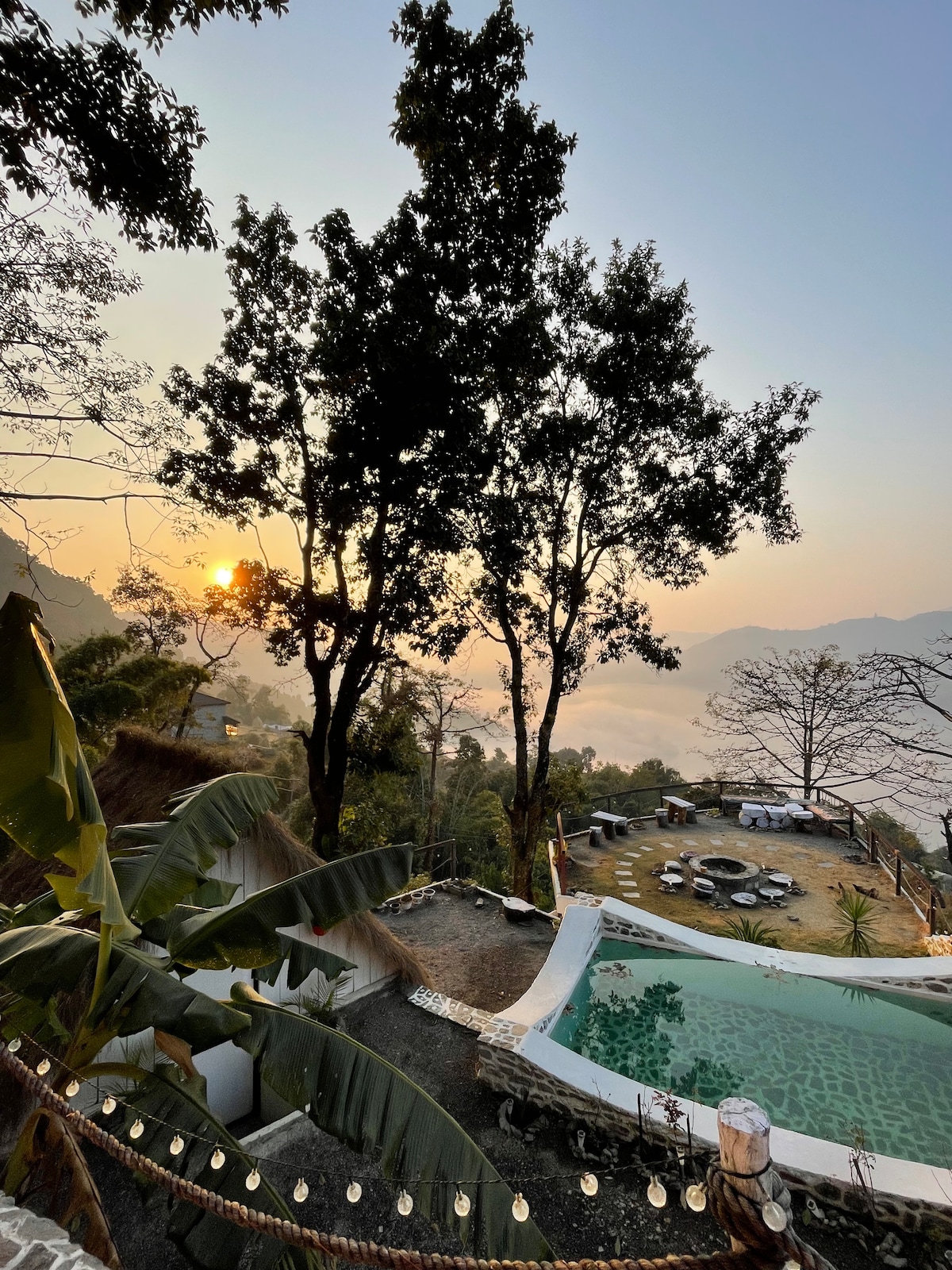 A serene view of a swimming pool is captured, surrounded by lush greenery and tall trees. In the distance, sunrise casts a warm glow over a scenic landscape. A gathering area with seating is visible, enhancing the peaceful outdoor setting.
