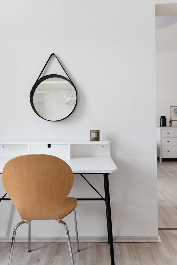A minimalist workspace is shown featuring a white desk with a small drawer and a wooden chair. A round black mirror hangs above the desk, reflecting the soft, neutral tones of the wall. A glimpse of the adjoining room is visible in the background.