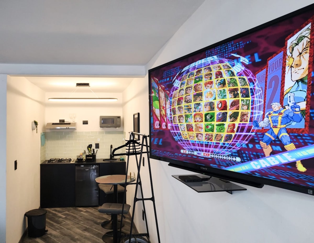 An open kitchen area is visible, featuring sleek appliances and a small dining table set for two. A large flat-screen television displays a classic video game menu. A coat rack stands nearby, and a trash bin completes the layout, highlighting a functional living space.