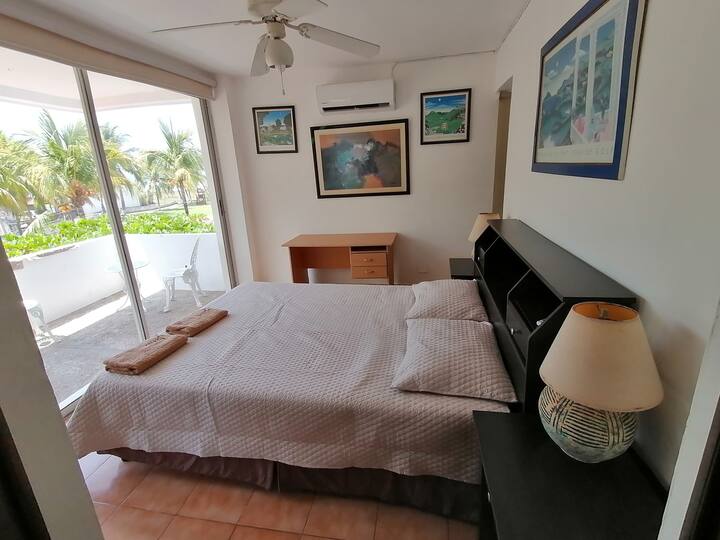 Habitación principal con cama tamaño queen, AC y ventilador de techo y vista a la terraza y jardin de cocos y al final la playa. 