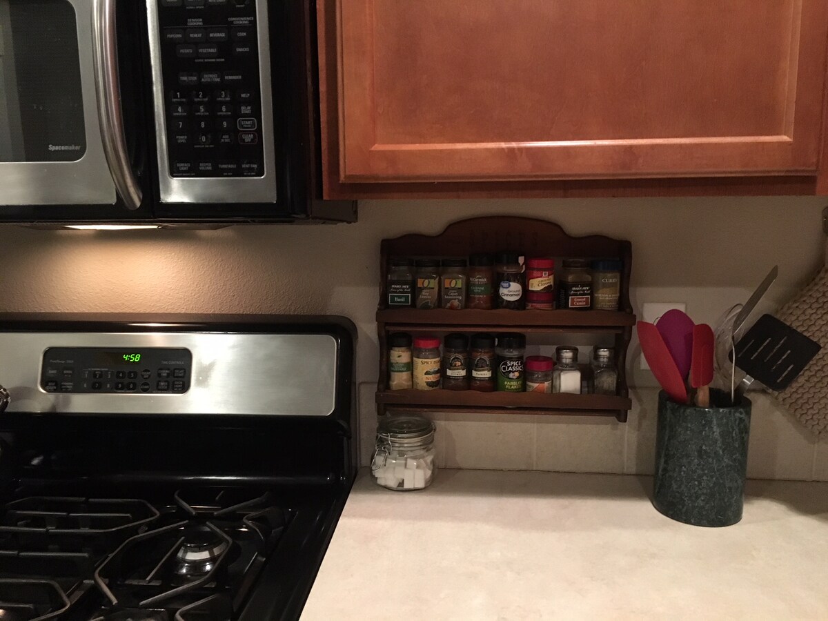 A well-organized kitchen counter displays a spice rack mounted on the wall, containing various spice jars. A stainless steel stove and microwave are visible alongside a set of utensils in a holder. A glass jar sits on the countertop, adding to the kitchen's functionality.