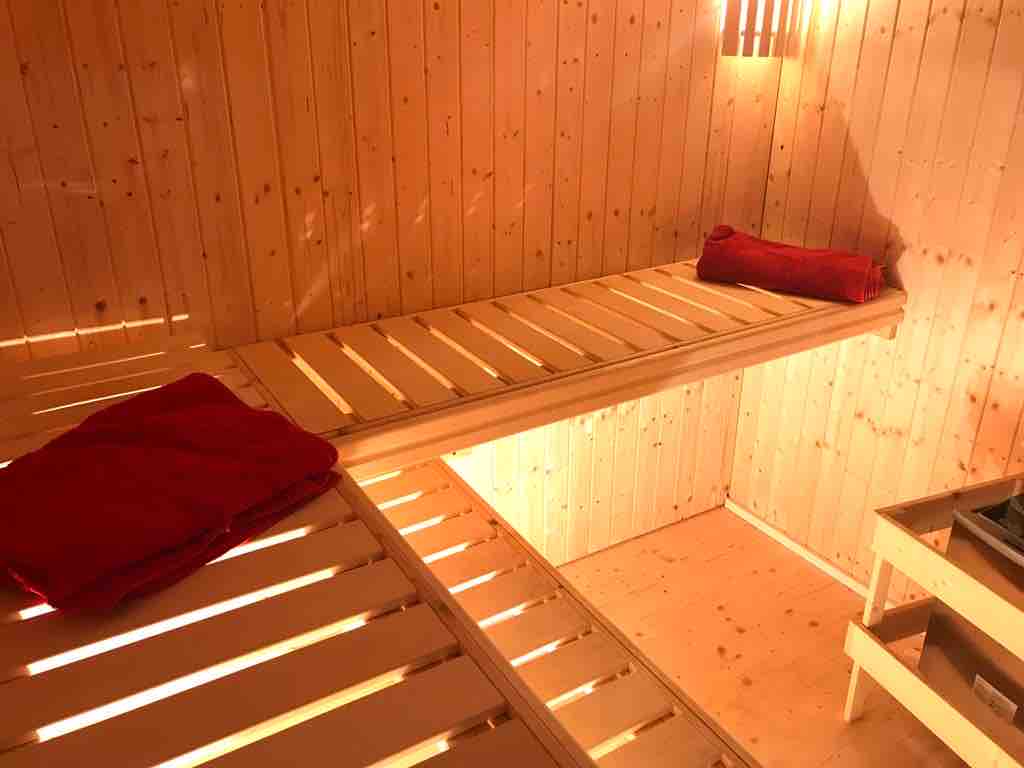A wooden sauna interior is displayed, featuring two levels of slatted benches made from light wood. Soft red towels are neatly placed on the benches. Ambient lighting creates a warm glow, highlighting the natural wood paneling surrounding the space.