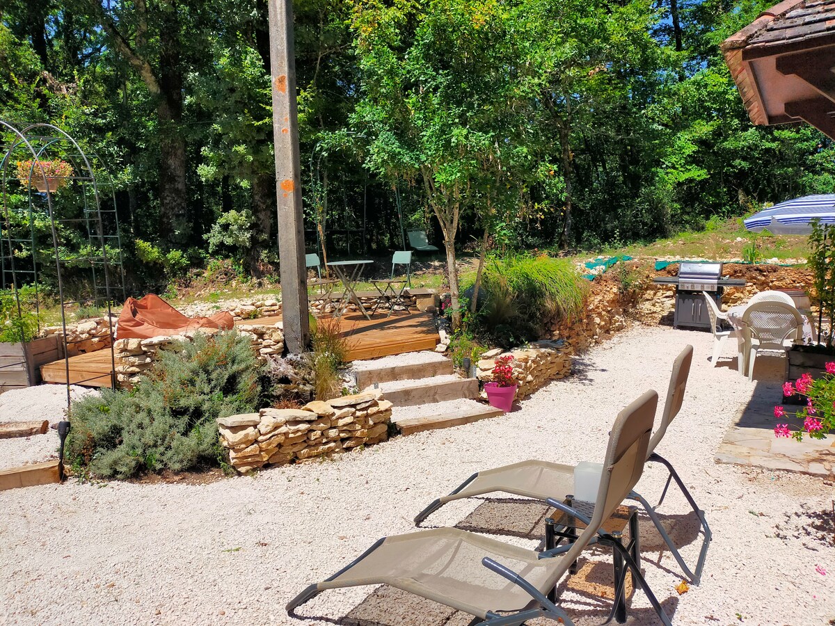 A serene outdoor space features lounge chairs positioned on gravel, surrounded by greenery and stone pathways. Tables and additional seating areas are visible, contributing to a peaceful ambiance. A shaded area with a grill is present, enhancing the outdoor gathering potential.