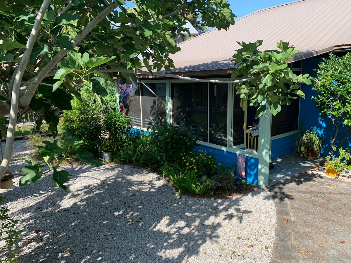 The exterior of the cottage is surrounded by lush greenery and colorful flowers, providing a welcoming atmosphere. A gravel pathway leads to the entrance, which features a screened porch and a vibrant blue facade. Trees offer shade and enhance the tranquil setting.