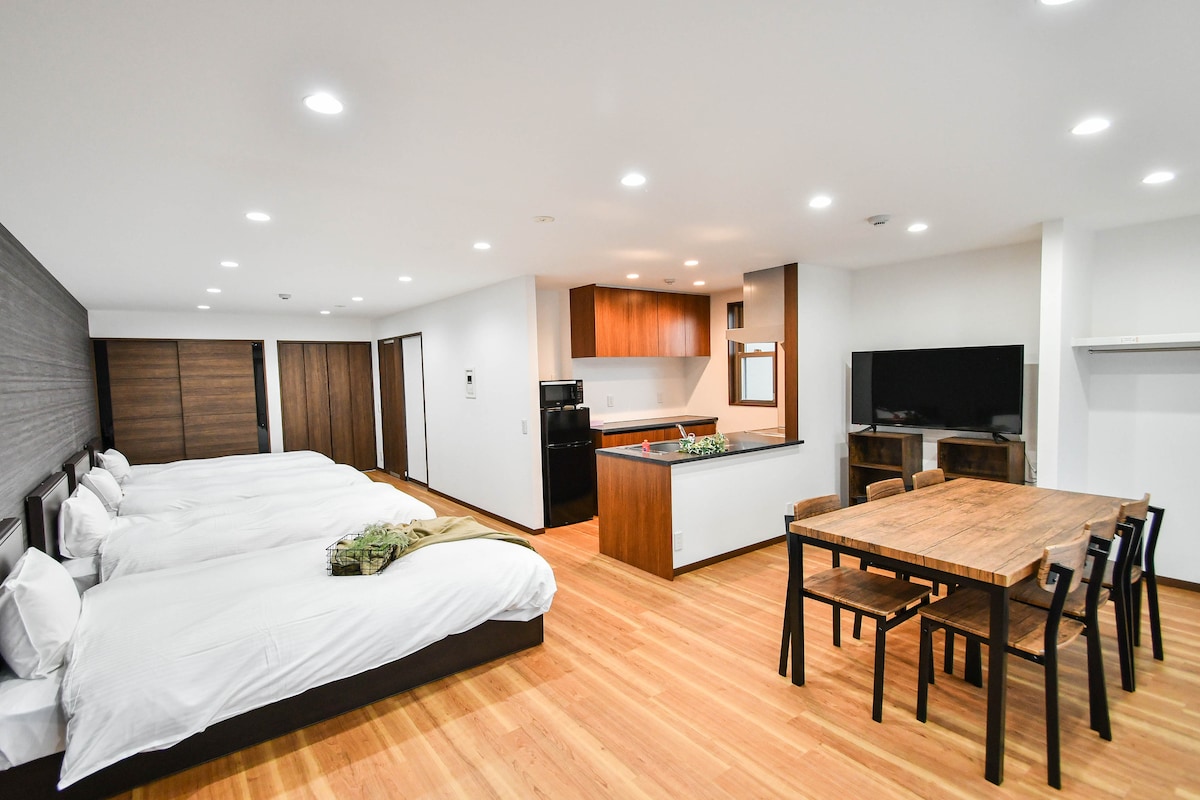 This spacious room features two sets of beds with soft white linens. An open kitchenette and dining area is highlighted by a wooden table surrounded by black chairs. A large television is mounted on the wall, and warm wooden floors enhance the inviting atmosphere.
