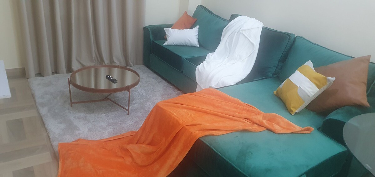 A spacious sitting area is furnished with a deep green sectional sofa and a round coffee table. An orange throw and white blanket drape casually over the sofa. Soft, neutral curtains frame the window, enhancing the warm aesthetic of the room.
