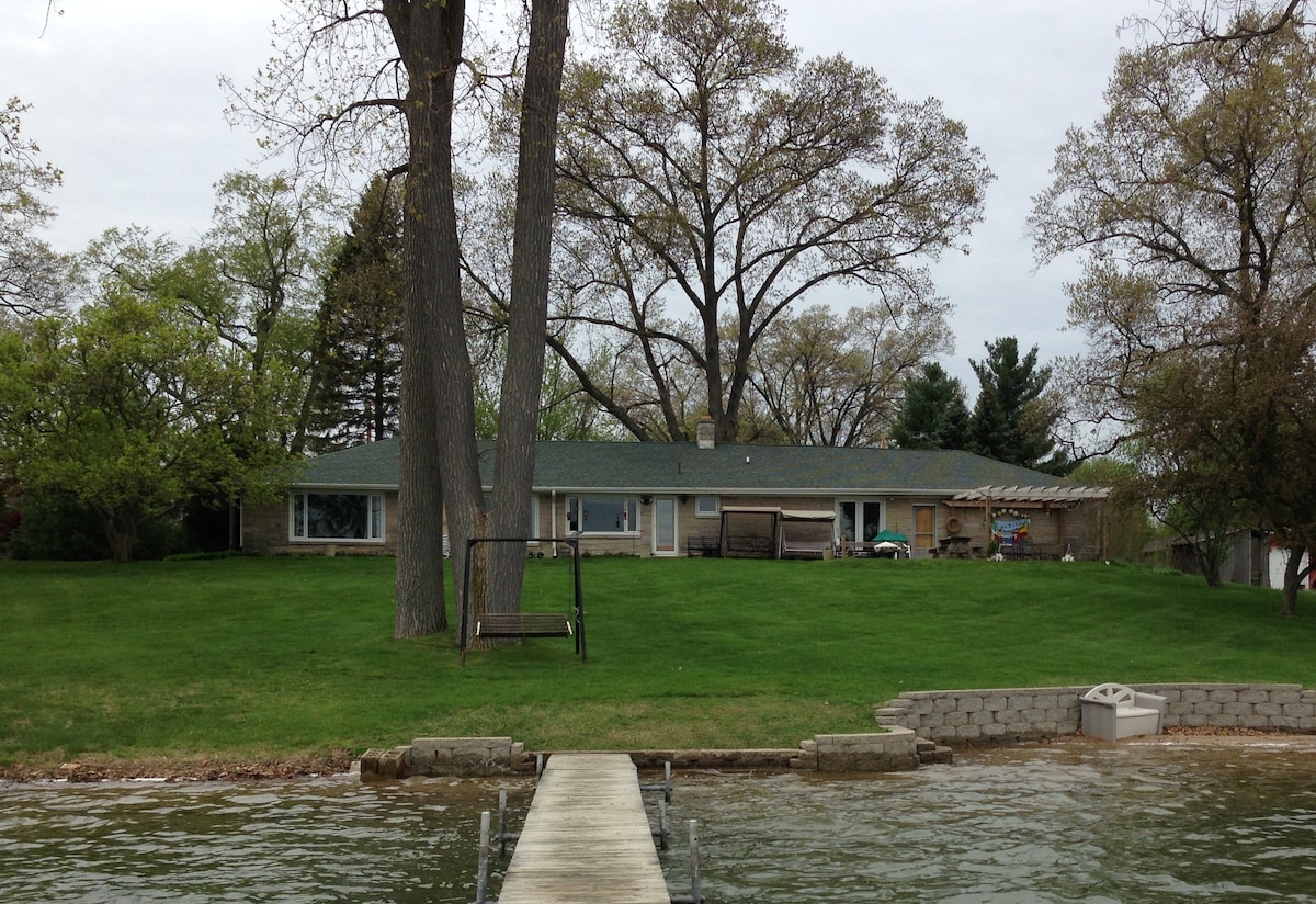 A lakehouse is visible from the water, featuring a spacious yard with green grass and mature trees. A wooden dock extends towards the house, which has large windows and a patio area. A swing can be seen in the yard, enhancing the natural setting.