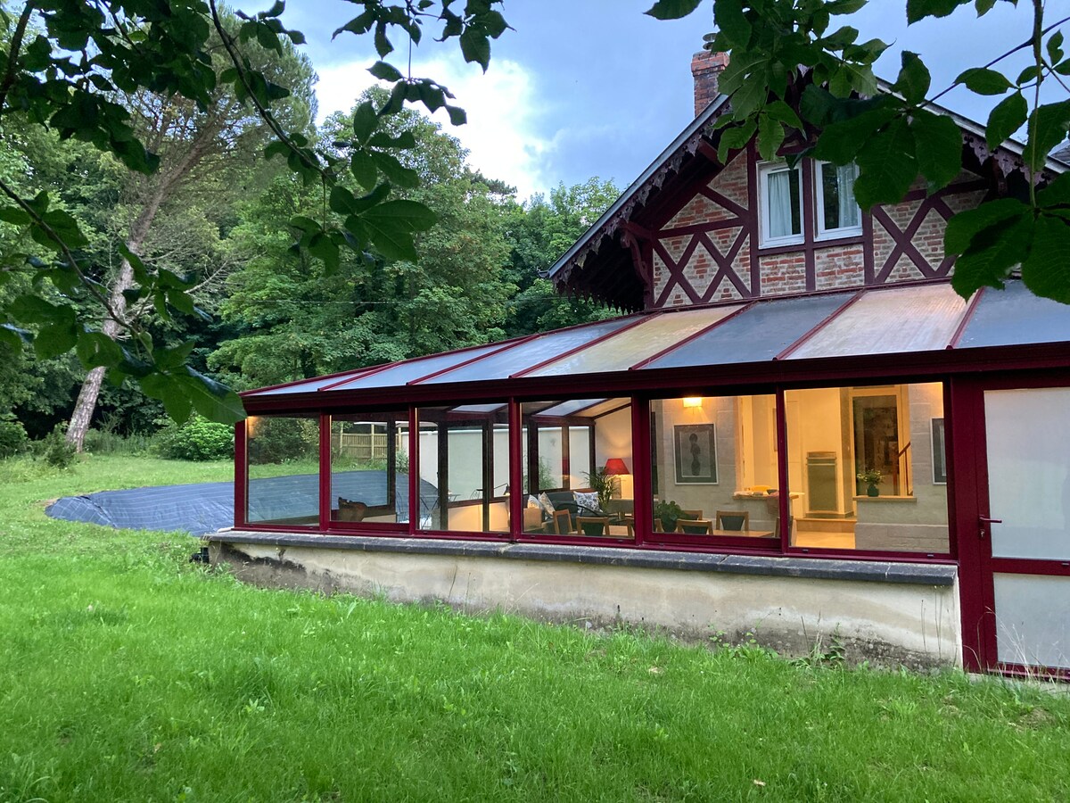 A charming house is framed by lush greenery, with a glass-walled sunroom showcasing warm indoor lighting. The scenic view includes a well-kept lawn and tree coverage, creating a tranquil retreat. The structure features a blend of traditional and modern architectural elements.