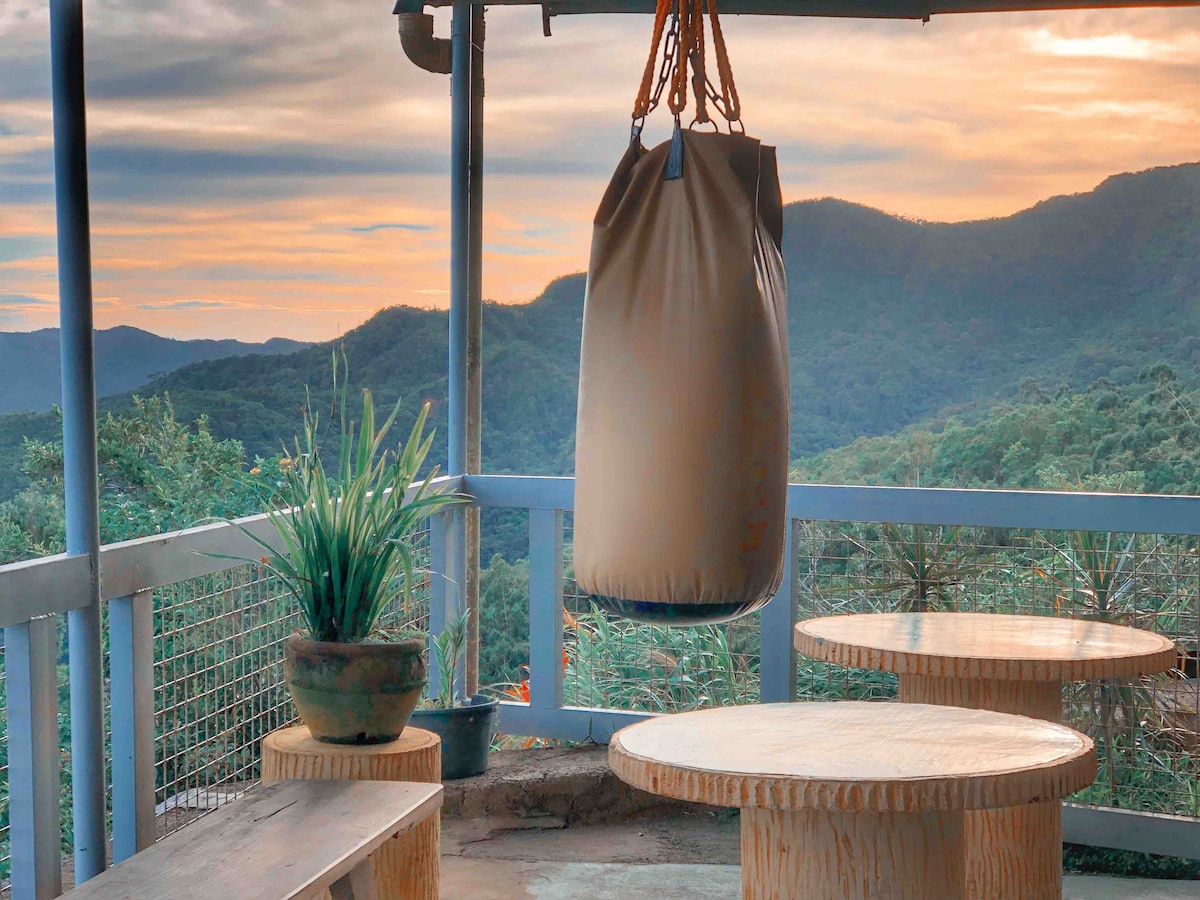 A serene outdoor area features a hanging bag, ideal for relaxation, surrounded by scenic mountain views. Two round wooden tables are placed on a textured surface, complemented by a potted plant. The evening sky casts a warm light over the landscape, enhancing the peaceful atmosphere.