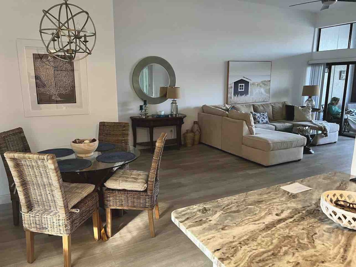 A dining area features a round glass table surrounded by four woven chairs. A beige sectional sofa is positioned against the wall, with a large round mirror above a side table. Sunlight enters through sliding glass doors, illuminating the neutral-toned space.