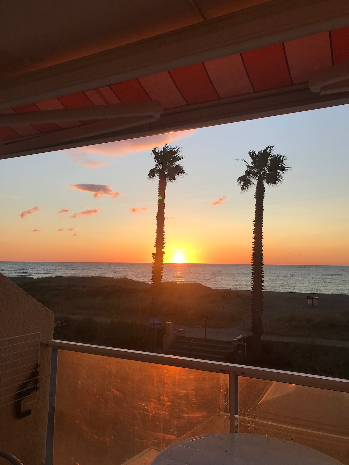The view of a tranquil sunset is framed by the balcony. Silhouetted palm trees stand against a vibrant sky with hues of orange and pink, while the ocean reflects the fading sunlight. The scene offers direct sightlines to the beach.