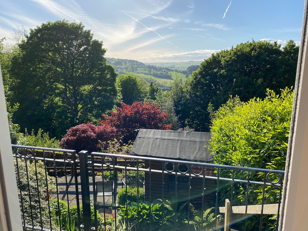 A view from the balcony showcases lush greenery and vibrant foliage, including trees and colorful shrubs. The distant hills are visible under a clear blue sky, creating a sense of tranquility and connection with nature.