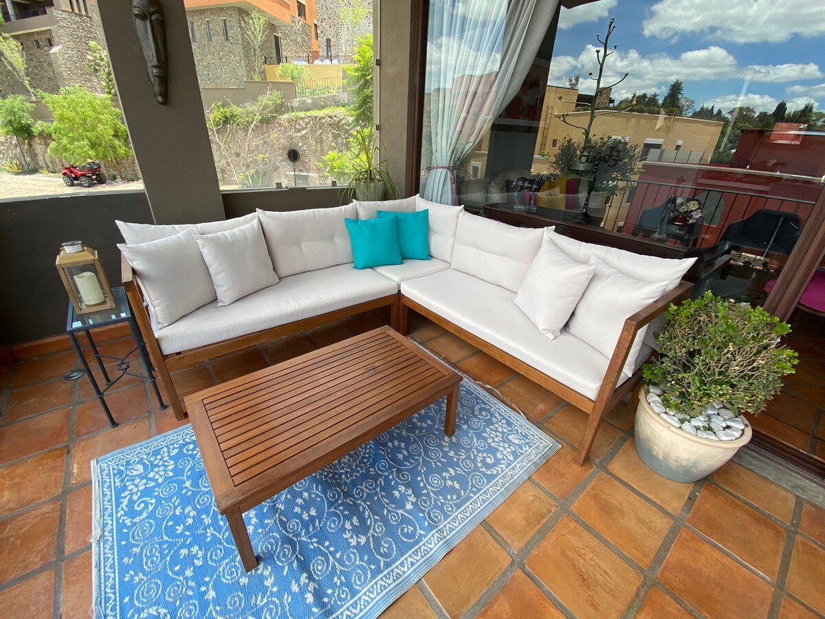 A comfortable outdoor seating area is furnished with a spacious white sectional sofa adorned with decorative pillows. A wooden coffee table is centered on a patterned area rug, while greenery and light curtains frame the view of the terrace.