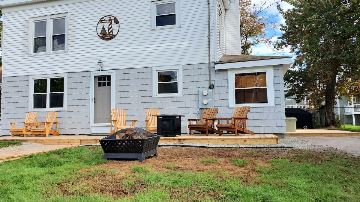 An outdoor patio area is presented with several wooden seating options arranged around a fire pit. The light-colored siding of the house features multiple windows, allowing natural light to enhance the welcoming space.