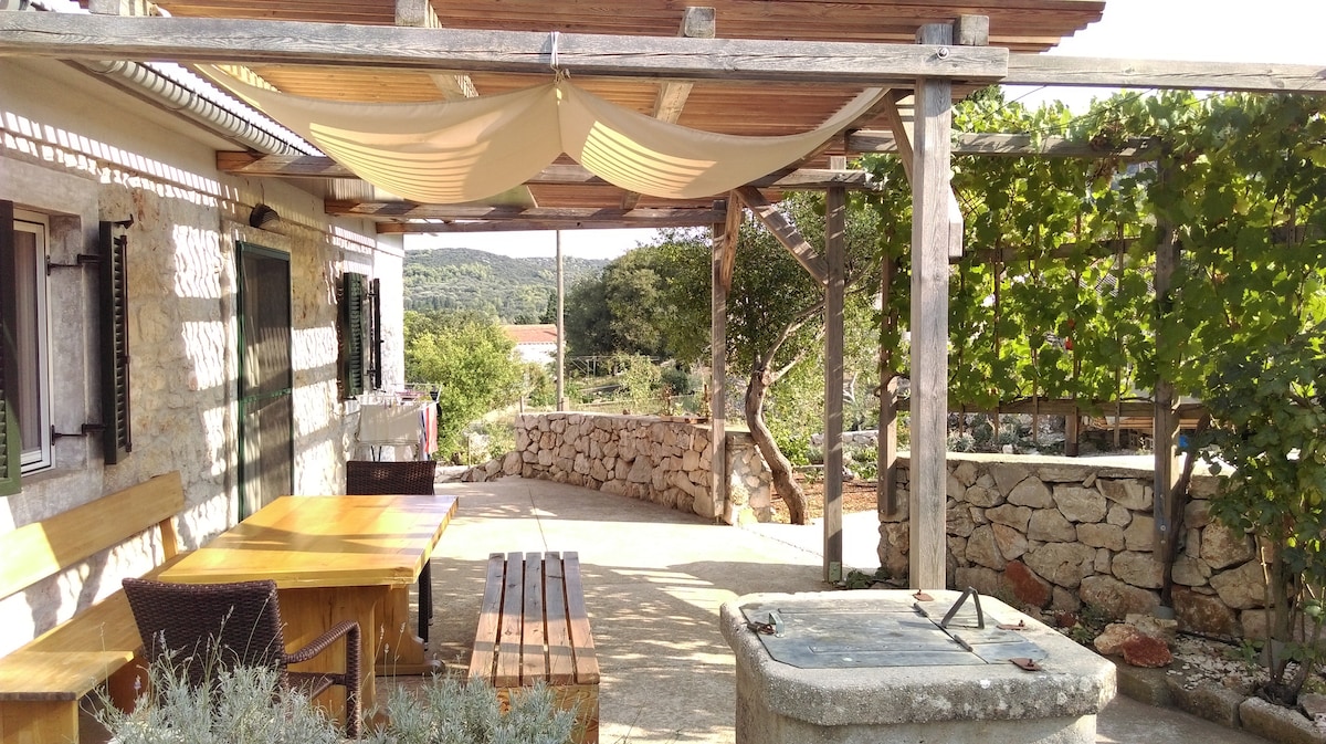 An outdoor terrace is shown, featuring a wooden dining table accompanied by chairs. A large, shaded area is created by fabric drapes overhead. Natural stone walls frame the space, while greenery and trees provide a serene backdrop, emphasizing the tranquil environment.