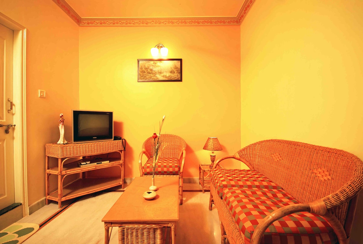 A cozy living area features two seating arrangements with woven furniture, each adorned with patterned cushions. A small wooden coffee table sits at the center, while a flat-screen television is positioned against the wall. Soft lighting is provided by a mounted fixture and a standing lamp.
