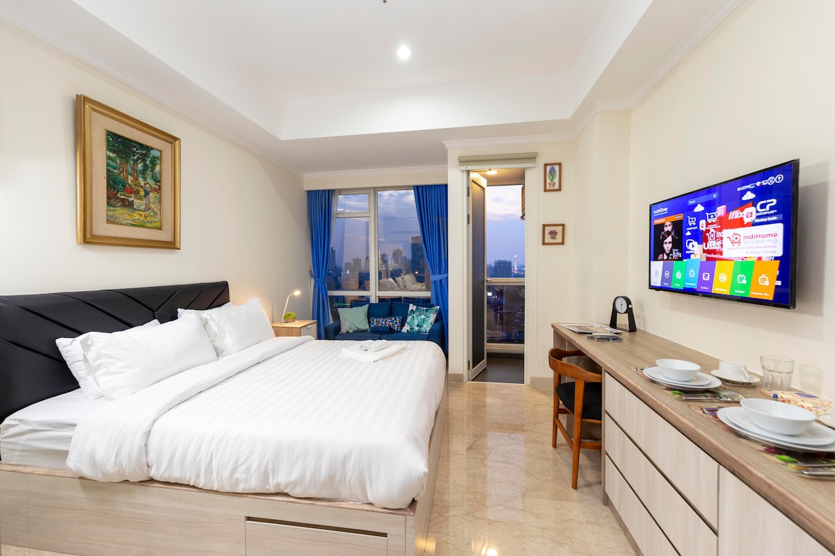 A studio room is furnished with a spacious bed, complemented by a bedside lamp and artwork on the wall. A Smart TV is mounted opposite the bed, and a functional desk with dining utensils is set nearby. Large windows provide views of the city skyline.