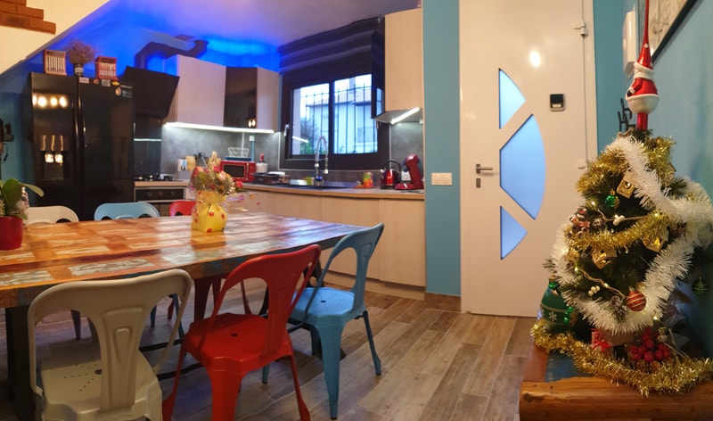 The kitchen area features a colorful dining table surrounded by red, blue, and white chairs. Modern appliances are showcased in a well-lit space, while a small Christmas tree decorates one corner, adding a festive touch to the vibrant environment.