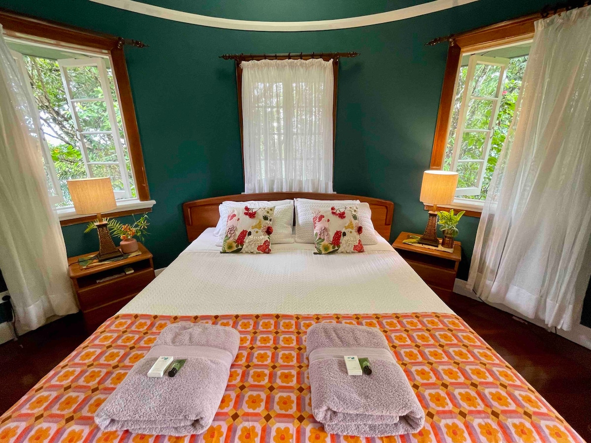The round bedroom features a spacious bed adorned with decorative pillows and a colorful throw. Soft lighting from two bedside lamps complements the green walls. Sheer curtains frame windows that offer views of the surrounding greenery.