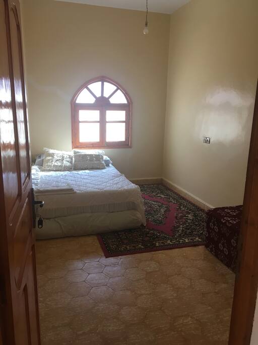 Habitación de invitados con cama de matrimonio perchero dos puuf, un perchero,puerta con llave, ventana con rejas y mosquiteros