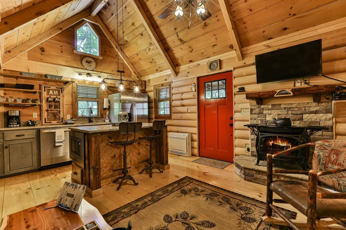 A rustic cabin interior features wooden beams and a cozy ambiance. The spacious kitchen includes modern appliances and a charming island with seating. A stone fireplace sits against one wall, while an inviting red door offers a pop of color, enhancing the warm, natural surroundings.
