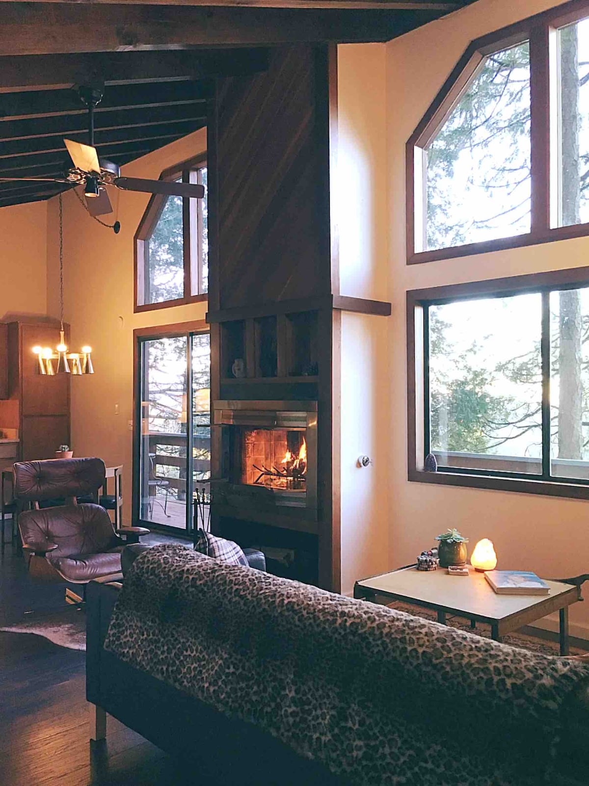 The spacious living area features large windows offering views of the surrounding forest. A cozy gas fireplace is positioned between natural wood accents. Leather chairs and a small wooden table provide a comfortable gathering space, complemented by soft lighting from a modern chandelier above.