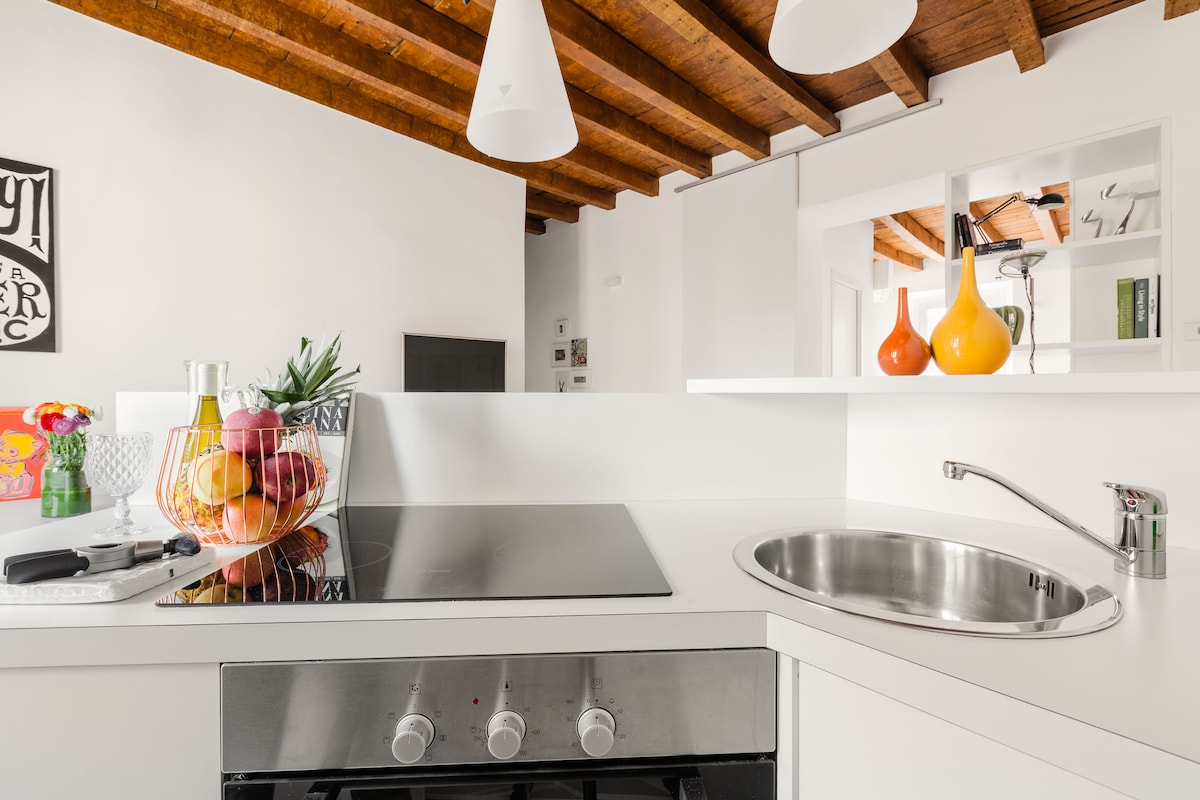 A modern kitchen features sleek white countertops and wooden ceiling beams. An induction cooktop and stainless-steel sink are visible. A bowl of fresh fruit sits prominently, accompanied by decorative glass vases in vibrant colors, enhancing the clean and functional design.