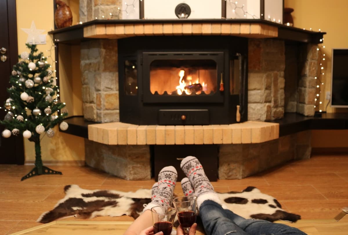 A cozy living area features a stone fireplace, with a warm fire burning inside. A small decorative Christmas tree is placed nearby, and two pairs of feet in fuzzy socks can be seen resting on a soft rug, holding glasses of a dark beverage.